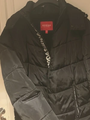 Svart pufferjacka från Guess - Svart pufferjacka från Guess med logotyp längs dragkedjan och på ärmen. Jackan har quiltad design, huva och två fickor med dragkedja. Perfekt för kyliga dagar och ger en snygg, sportig look.