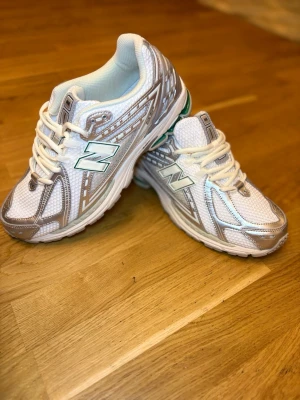 Vita och silver New Balance sneakers - Snygga vita sneakers från New Balance med detaljer i silver och meshmaterial för extra andningsförmåga. Klassisk logga på sidan och bekväm platt sula. Perfekta för dig som gillar sportig och trendig stil.
