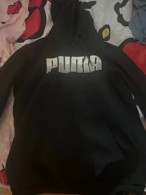 Svart hoodie från Puma med tryck - Snygg svart hoodie från Puma med stort vitt logotyptryck på bröstet. Klassisk huva med dragsko och känguruficka framtill. Perfekt för en avslappnad och sportig stil. Tillverkad i mjukt material för extra komfort.