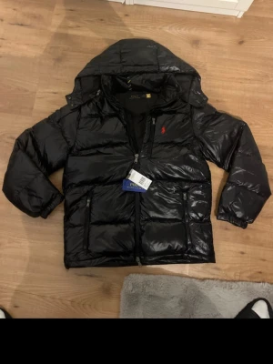 Svart dunjacka från Polo Ralph Lauren helt ny St M - Snygg svart pufferjacka från Polo Ralph Lauren med röd logga på bröstet. Jackan har huva, dragkedja framtill och två sidofickor med dragkedja. Perfekt för kalla dagar och riktigt stilren look. St M mvh Alexander 