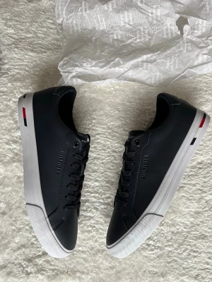 Svarta sneakers från Tommy Hilfiger - Snygga svarta sneakers från Tommy Hilfiger med vita sulor och diskreta logodetaljer på sidan och hälen. Skorna har klassisk snörning och är tillverkade i slätt syntetmaterial. Perfekta för en clean och stilren look. Skorna är köpta i USA har aldrig använt dem elr fått chansen o göra det därför säljer jag dem dem kostar 1600 nya kvitton finns