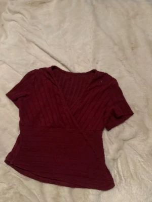 Vinröd omlottopp med ribbad struktur - Snygg vinröd omlottopp med ribbad struktur och korta ärmar. Toppen har en v-ringad design och sitter figurnära. Perfekt att matcha med jeans eller kjol för en trendig look.