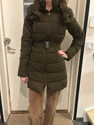 Gant-Mörkgrön dunjacka med bälte - Gant, Lång, mörkgrön dunjacka med quiltad design och avtagbart bälte i midjan. Jackan har kapuschong/som blir som hög krage och som går att ta bort. Modellen är 175cm och 34/36