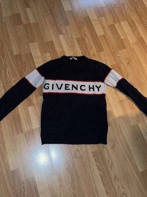 Svart stickad tröja från Givenchy - Snygg svart stickad tröja från Givenchy med vit panel och röd kant över bröstet där loggan står i stora bokstäver. Långa ärmar med vita och röda detaljer. Klassisk rund halsringning och ribbade muddar. Perfekt för dig som gillar exklusiva streetwear-plagg.