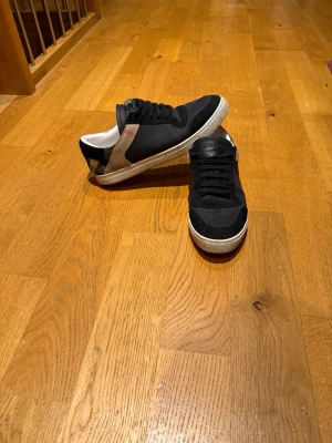 Svarta Burberry sneakers med rutiga detaljer - Snygga svarta sneakers från Burberry med klassiska rutiga detaljer på sidan och vit sula. Skorna har snörning och en rund tå. Perfekta för dig som vill ha en stilren och tidlös look med en touch av lyx.