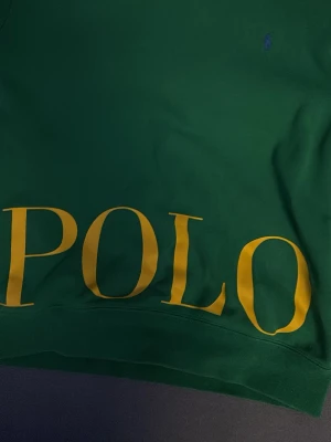 Grön hoodie från Polo Ralph Lauren - Snygg grön hoodie från Polo Ralph Lauren med stor gul POLO-text framtill och diskret logga på bröstet. Tillverkad i mjuk bomullsblandning som känns skön mot huden. Perfekt för dig som vill sticka ut med klassisk streetwear-vibe.