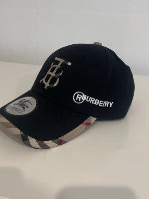 Svart keps med Burberry-inspirerad skärm - Snygg svart keps med broderad logga framtill och texten BRURBERRY' på sidan. Skärmen har ett rutigt mönster i beige, svart och rött som påminner om Burberrys klassiska stil. Justerbar passform och cool streetwear-vibe.