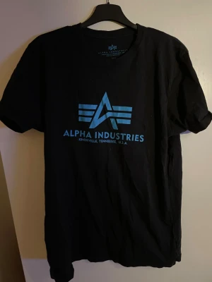 Svart Alpha Industries t-shirt - Svart t-shirt från Alpha Industries med blå logga och text framtill. Klassisk rund halsringning och korta ärmar. Perfekt för en avslappnad och cool stil. Tillverkad i mjuk bomull för skön komfort.