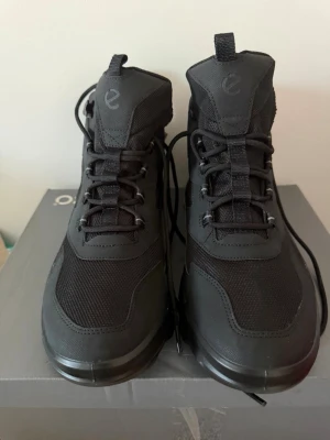 Svarta kängor från Ecco MX - Säljer ett par svarta Ecco MX kängor med snörning och robust sula. Tillverkade i en mix av skinn och syntetmaterial, med meshpaneler för extra andningsförmåga. Perfekta för dig som vill ha stilrena och funktionella boots till vardags.