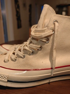 Converse nyskick  - Klassiska vita höga sneakers i canvas med snörning och röd rand längs sulan. Skorna har rund tå, platt sula och metallhål för snörena. Perfekta för en avslappnad och stilren look.