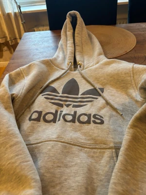 Grå hoodie från Adidas Originals - Snygg grå hoodie från Adidas Originals med stor svart Trefoil-logga och text på bröstet. Klassisk känguruficka framtill och justerbar huva med dragsko. Tillverkad i mjukt sweatshirtmaterial som är perfekt för chill dagar.