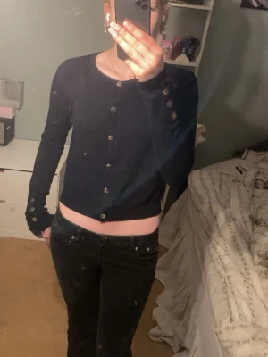 Mörkblå cardigan med guldknappar - Snygg mörkblå cardigan med guldknappar framtill och på ärmsluten. Tröjan har en figurnära passform, rund halsringning och långa ärmar. Perfekt att styla med jeans eller kjol för en trendig look.