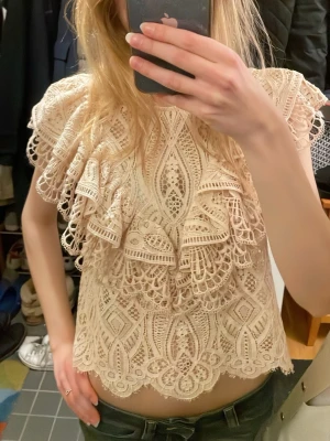 Beige spetsblus med volanger från Zara - Supersöt beige topp/blus från Zara i spets med detaljerade broderier och volanger framtill. Kort ärm och croppad passform, perfekt till både jeans och kjol. Blusen har rund halsringning och knäppning bak i nacken. Köpt för ett tag sedan men använd ett fåtal gånger. 💓