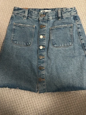 Blå jeanskjol från Zara Trafaluc - Säljer en blå jeanskjol från Zara Trafaluc med stora fickor fram och bak, knappar längs hela framsidan och rå kant nertill. Kjolen har bälteshällor och en klassisk A-linjeformad passform. Perfekt till sommaren!