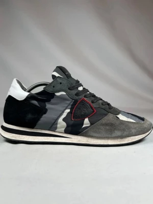 Philippe Model sneakers i camo - Snygga Philippe Model sneakers med svart, grå och vit camouflage-mönster. Skorna har detaljer i mocka, snörning och en röd kant runt loggan på sidan. Platt sula och rund tå för en klassisk streetwear-look.