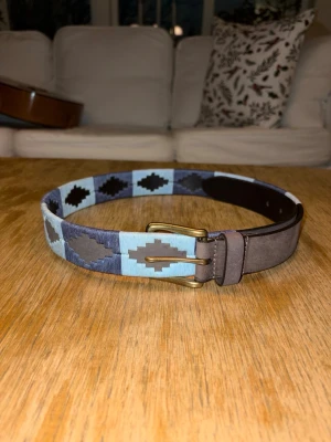 Navy blue nordfors bälte - Snyggt bälte med blå och bruna geometriska mönster och mässingsfärgat spänne. Bältet har detaljer i brunt läder och passar perfekt för att ge din outfit en unik touch. Materialet är en mix av tyg och läder. Finns i storlek 80.90. 100 cm