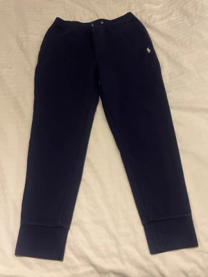 Mörkblå fleecebyxor från Polo Ralph Lauren - Mörkblå mjukisbyxor från Polo Ralph Lauren med broderad logga på vänster sida. Tillverkade i mjuk fleece, har breda muddar vid bensluten och en bakficka. Perfekta för chill dagar eller när du vill vara extra bekväm.