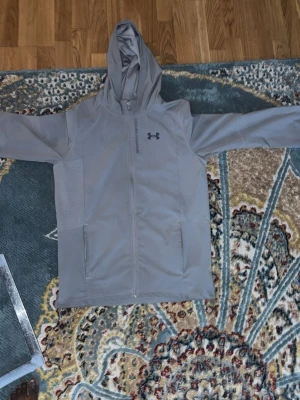 Grå vindjacka från Under Armour - Snygg grå vindjacka från Under Armour med huva och hel dragkedja framtill. Jackan har två sidofickor och en stilren logga på bröstet. Perfekt för blåsiga dagar och sportiga outfits. Materialet känns lätt och smidigt, och jackan har en modern passform. Priset kan fiskuteras.