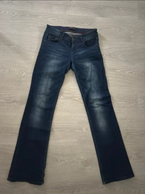 Mörkblå bootcut jeans med dragkedjedetalj - Snygga mörkblå bootcut jeans med cool dragkedjedetalj på fickan. Jeansen har klassisk femficksdesign och subtila slitningar framtill för en avslappnad look. Tillverkade i mjukt bomullsmaterial med en skön passform.