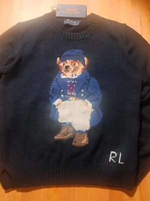 Mörkblå stickad tröja Polo Bear - Mörkblå stickad tröja från Polo Ralph Lauren med ikoniska Polo Bear-motivet framtill. Tröjan har rund hals, ribbade muddar och RL-broderi nedtill. Perfekt för dig som vill ha en klassisk men lekfull look. Sitter som S/XS