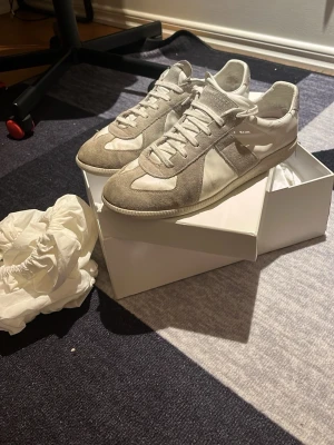 Maison Margiela (äkta) - Snygga sneakers från Maison Margiela i vitt och ljusgrått. Skorna har klassisk design med rund tå, snörning och paneler i mocka och skinn. Perfekta för dig som gillar stilrena och trendiga sneakers med exklusiv känsla.