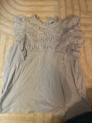 Ljusblå blus med spets från Holly & Whyte - Supersöt ljusblå blus från Holly & Whyte by Lindex med broderade spetsdetaljer och volanger upptill.  Aldrig använd.