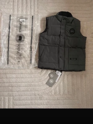 Mörkgrå dunväst från Canada Goose - Snygg mörkgrå dunväst från Canada Goose med klassisk logga på bröstet och tryckknappar framtill. Västen har hög krage och två praktiska fickor. Tillverkad i slitstarkt material och fylld med dun för extra värme. Perfekt för lager-på-lager och streetwear. Str m helt ny 