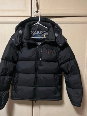 Svart dunjacka från Polo Ralph Lauren - Snygg svart dunjacka från Polo Ralph Lauren med huva och dragkedja framtill. Jackan har en quiltad design, två sidofickor med dragkedja och en liten röd logga på bröstet. Perfekt för kalla dagar och riktigt stilren look.