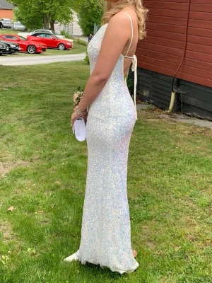 Glittrig vit långklänning med snörning - Supersnygg vit långklänning med tunna axelband och öppen rygg med snörning. Klänningen är täckt av glittriga paljetter som ger ett magiskt skimmer. Perfekt för bal eller student! Materialet känns mjukt och följsamt. 