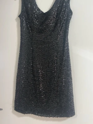 Svart glittrig paljettklänning - Säljer en svart kort klänning täckt med glittrande paljetter. Klänningen har en djup v-ringning och är ärmlös, perfekt för dig som vill sticka ut. Materialet känns festligt och skimrar snyggt i ljuset.