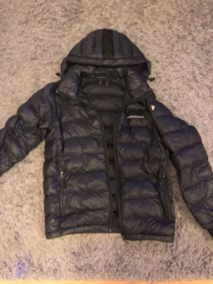 Svart pufferjacka Peak Performance - Säljer en svart pufferjacka från Peak Performance med huva och dragkedja. Jackan har två sidofickor med dragkedja och diskret logga på bröstet. Perfekt för kalla dagar och riktigt skön att bära.