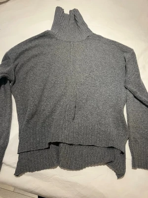 Grå polotröja med ribbad krage - Mysig grå polotröja med hög, ribbad krage och lång ärm. Tröjan har en avslappnad passform och ribbade muddar vid ärmslut och nederkant. Perfekt för kyliga dagar och enkel att styla med jeans eller kjol.