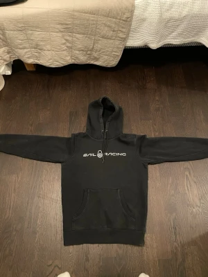 Svart hoodie från Sail Racing - Svart hoodie från Sail Racing med stor ficka framtill och vit logga tryckt över bröstet. Klassisk huva med dragsko och mjukt, borstat innerfoder för extra komfort. Perfekt för en avslappnad och sportig stil.