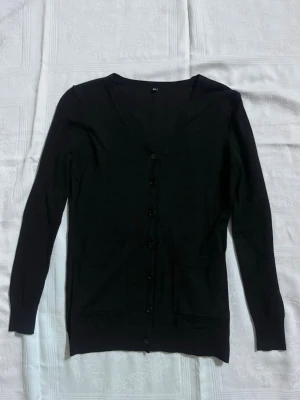 Svart kofta med knappar - En klassisk svart kofta med v-ringning och knäppning framtill. Tröjan har två fickor fram och är långärmad. Perfekt att slänga över en t-shirt eller linne för en chill look.