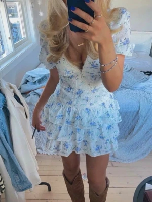 Blommig kortklänning med volanger - Supersöt kortklänning i vitt med blått blommönster och volanger. Klänningen har v-ringning, puffiga korta ärmar och flera lager volang i kjolen. Perfekt för sommarens alla tillfällen och ger en romantisk vibe.