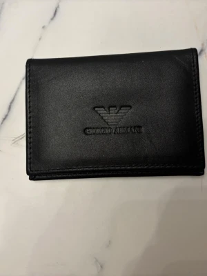 Svart plånbok från Giorgio Armani - Stilren svart plånbok från Giorgio Armani i skinn med präglad logga på både utsidan och insidan. Plånboken har flera kortfack och ett fönsterfack för ID eller körkort. Perfekt storlek för fickan och enkel att ta med.