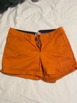 Orangea shorts i bomull - Snygga orangea shorts med uppvikta benslut och klassisk knapp- och dragkedjestängning. Shortsen har bälteshällor, sidofickor fram och två dekorativa fickor bak. Perfekta för varma dagar och ger en färgklick till din outfit.