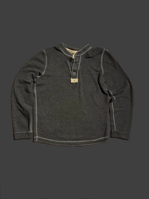 Mörkgrå vintage Hollister långärmad topp med knappar - Mysig mörkgrå långärmad topp med kontrastsömmar och knappdetaljer framtill. Tillverkad i mjukt bomullsmaterial, perfekt för lager på lager. Enkel och stilren design som passar till det mesta.