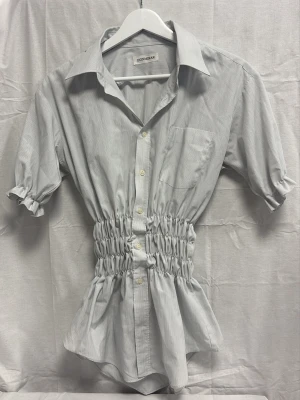 Vit skjorta med ljusblå ränder och smockad midja - Vit skjorta från DONMIRAR med smala blå ränder, smockad midja och ärmar. Klassisk krage, bröstficka och knappar framtill. Det finns tyvärr en fläck innanför kragen som vi tror kommer från en klisterlapp, fläcken är ej synlig när skjortan är på! Storleken står ej på skjortan men vi tror att den passar dig som bär storlek M och S (för en oversized passform).