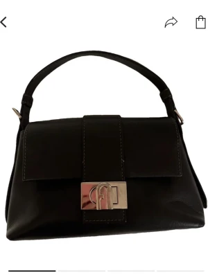 Svart handväska i skinn från Furla - AI beskrivning idk Stilren svart handväska från Furla i äkta skinn med elegant silverfärgat spänne framtill. Väskan har ett klassiskt, avlångt format och ett kort handtag som gör den enkel att bära i handen eller på armen. Perfekt accessoar för en trendig look.