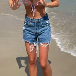 Blå denimshorts med fransar - Säljer ett par blå denimshorts med hög midja och slitna detaljer. Shortsen har fransig kant nedtill och klassisk femficksdesign. Perfekta för sommaren och ger en avslappnad look.