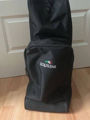 Svart ryggsäck från Equiline - Rymlig svart ryggsäck från Equiline med broderad logga och italiensk flagga på framsidan. Väskan har flera fack med dragkedja och bred axelrem med Equiline-tryck. Tillverkad i slitstark polyester, perfekt för dig som behöver en väska i stallet 