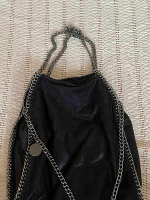 Svart handväska med kedjedetaljer - Snygg svart handväska från gina med glittrig finish och kraftiga silverfärgade kedjor runt kanterna och som axelrem. Väskan har en rund metallbricka med logga och en mjuk, slouchy form. Perfekt för dig som gillar statement-accessoarer.