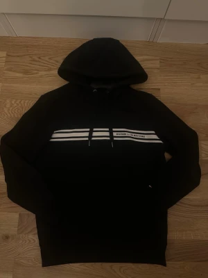 Svart hoodie från Tommy Hilfiger - Snygg svart hoodie från Tommy Hilfiger med vita ränder och texten 'HILFIGER Est. 1985 NEW YORK' över bröstet. Klassisk huva med dragsko och mjukt material som känns skönt mot huden. Perfekt för en avslappnad och trendig look.