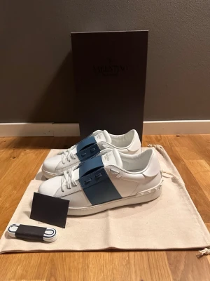 Valentino Garavani vita sneakers - Säljer ett par vita Valentino Garavani sneakers med bred blå rem över snörningen och subtila nitar på hälen. Skorna har rund tå, platt sula och är tillverkade i skinn. Klassisk och stilren design med logotyp på plösen.