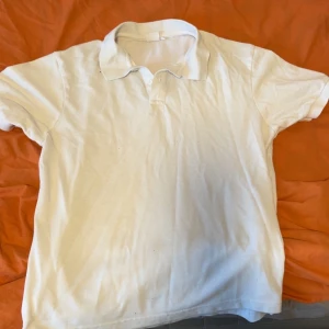 Vit pikétröja med krage - Klassisk vit pikétröja med kort ärm och krage. Tröjan har knappar vid halsen och är tillverkad i ett mjukt material som känns bekvämt mot huden. Perfekt till jeans eller shorts för en enkel och stilren look.