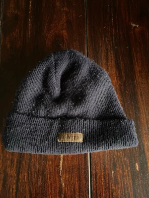 Mörkblå ribbstickad mössa Pointer - Mörkblå ribbstickad mössa från Pointer med uppvikt kant och en brun läderpatch med logga framtill. Klassisk modell som passar perfekt till kalla dagar. Materialet är mjukt och värmande, perfekt för höst och vinter.