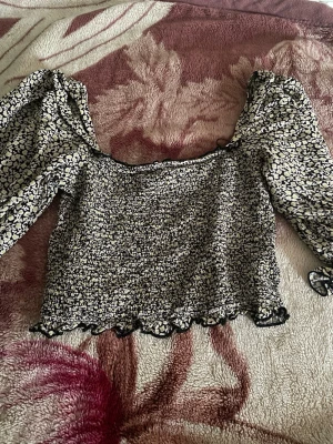 Blommig croppad blus med puffärm - Trendig croppad blus med svartvitt blommönster och puffiga långärmar. Toppen har smockad kropp, volangkant nertill och en söt, feminin look. Perfekt för dig som gillar statement-ärmar och romantisk stil.