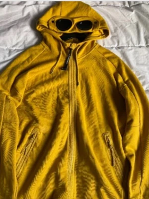 Gul hoodie med glasögon från C.P. Company - Säljer en gul hoodie från C.P. Company med hel dragkedja, ikoniska inbyggda glasögon i huvan och två fickor med dragkedja. Tillverkad i mjuk bomull för skön känsla och cool look. Perfekt för dig som vill sticka ut med streetwear-vibe.