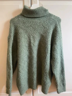 Grön stickad polotröja - Mysig grön stickad polotröja med strukturmönster och hög krage. Tröjan har lång ärm och avslappnad passform, perfekt för kalla dagar. Materialet ser ut att vara mjukt och värmande, troligen ullblandning.